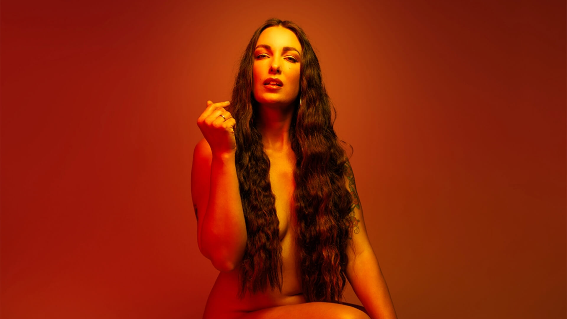 Frankie van Kan posing naked on an orange background. Her shoulder-length hair covering her body for Melt Festival 2024.