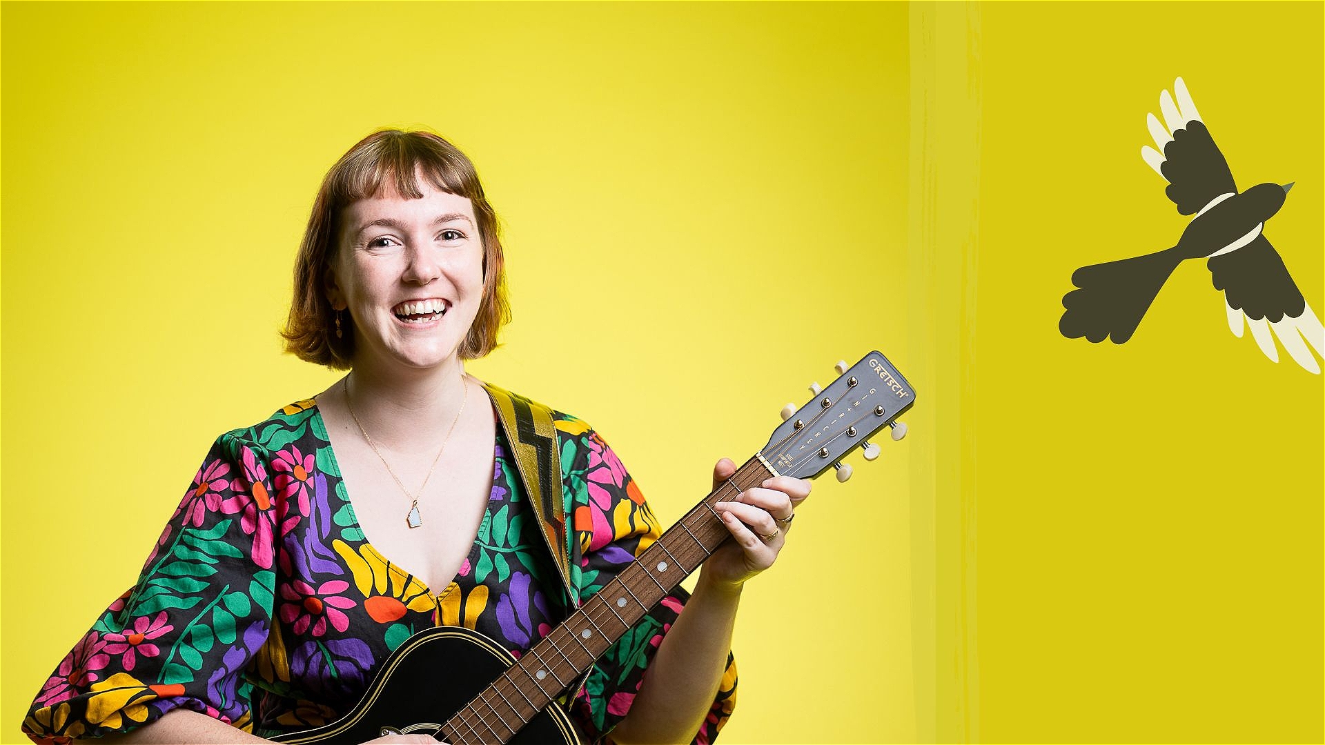 A person with short brown hair is smiling while playing an acoustic guitar. They are wearing a colorful, floral-patterned shirt. The background is bright yellow with a black and white bird silhouette on the right side.