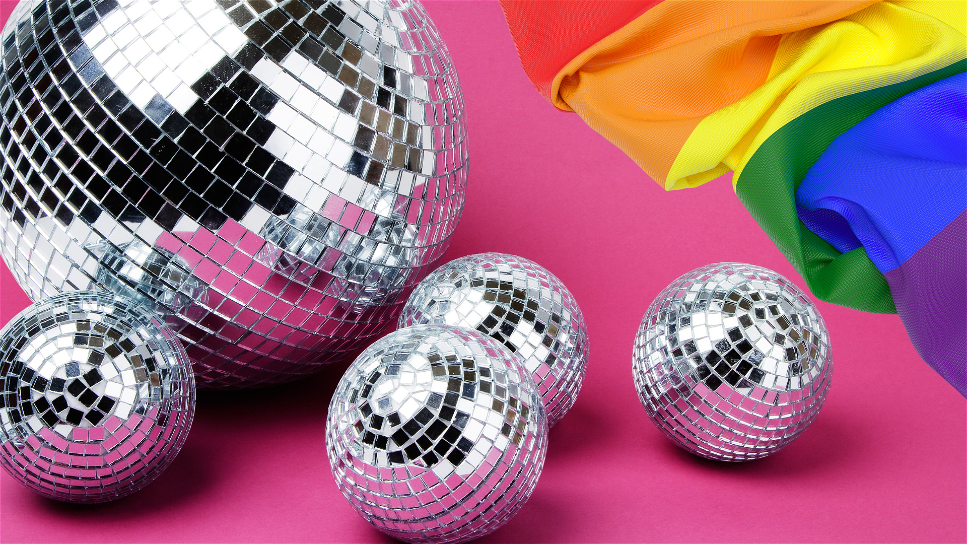 A composition with four small disco balls and one larger disco ball resting against a pink background. In the upper right, a vibrant rainbow-colored fabric is partially visible, adding a burst of color to the scene.