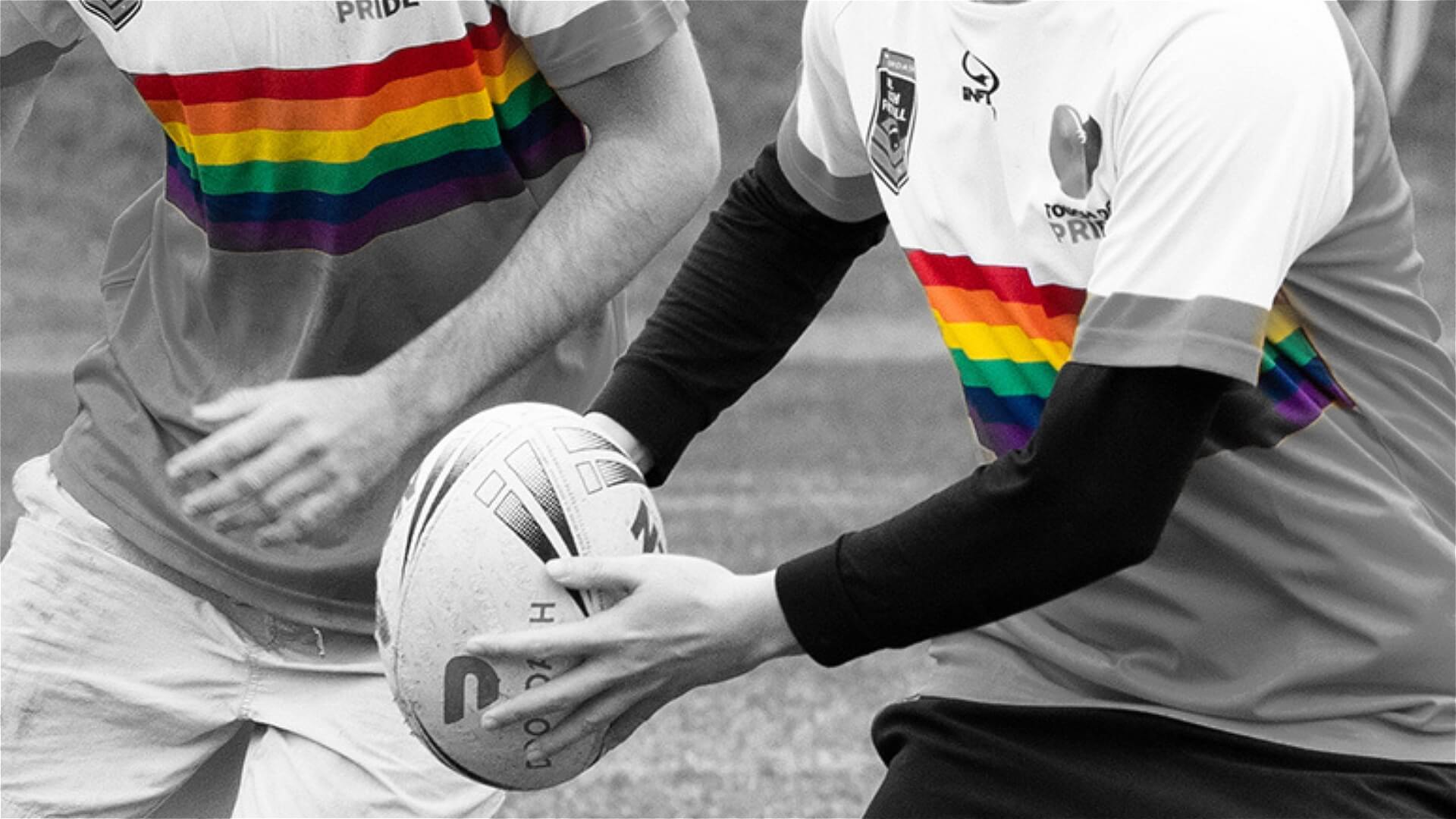 Two people in rugby uniforms with rainbow stripes, one holding a rugby ball, engage in a game. The photo is black and white except for the rainbow stripes on their shirts, emphasizing inclusion and diversity in sports.