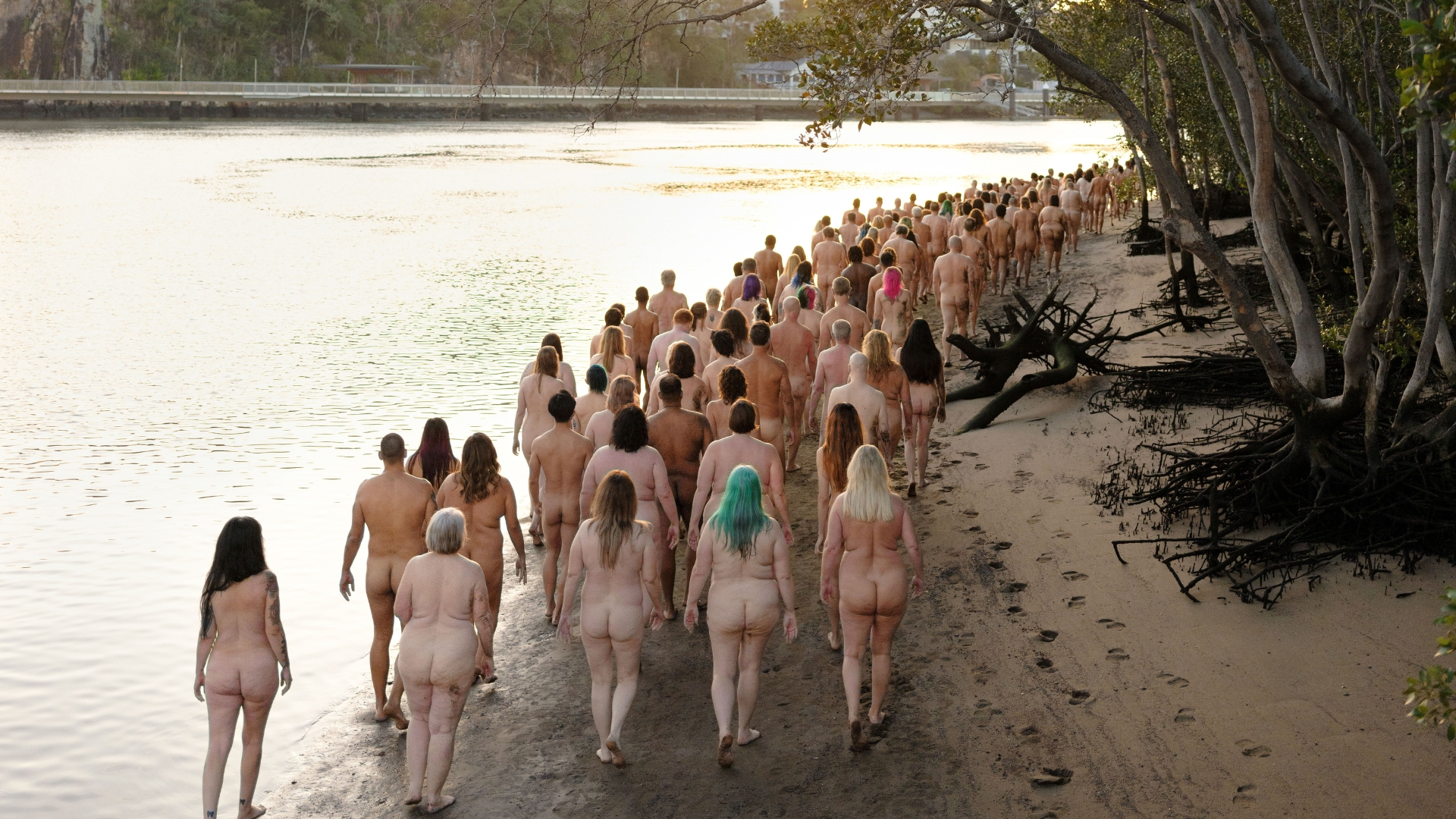 A large group of nude individuals walking along a sandy beach beside a body of water. The scene is set during daylight, with trees and footprints visible in the sand. The group is walking towards the distance, away from the camera.